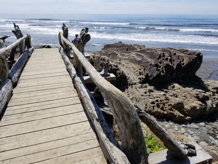 A wooden footbridge with artistically-hewn railings ends at a large folded rock. The ocean waves are in the background. It is sunny.